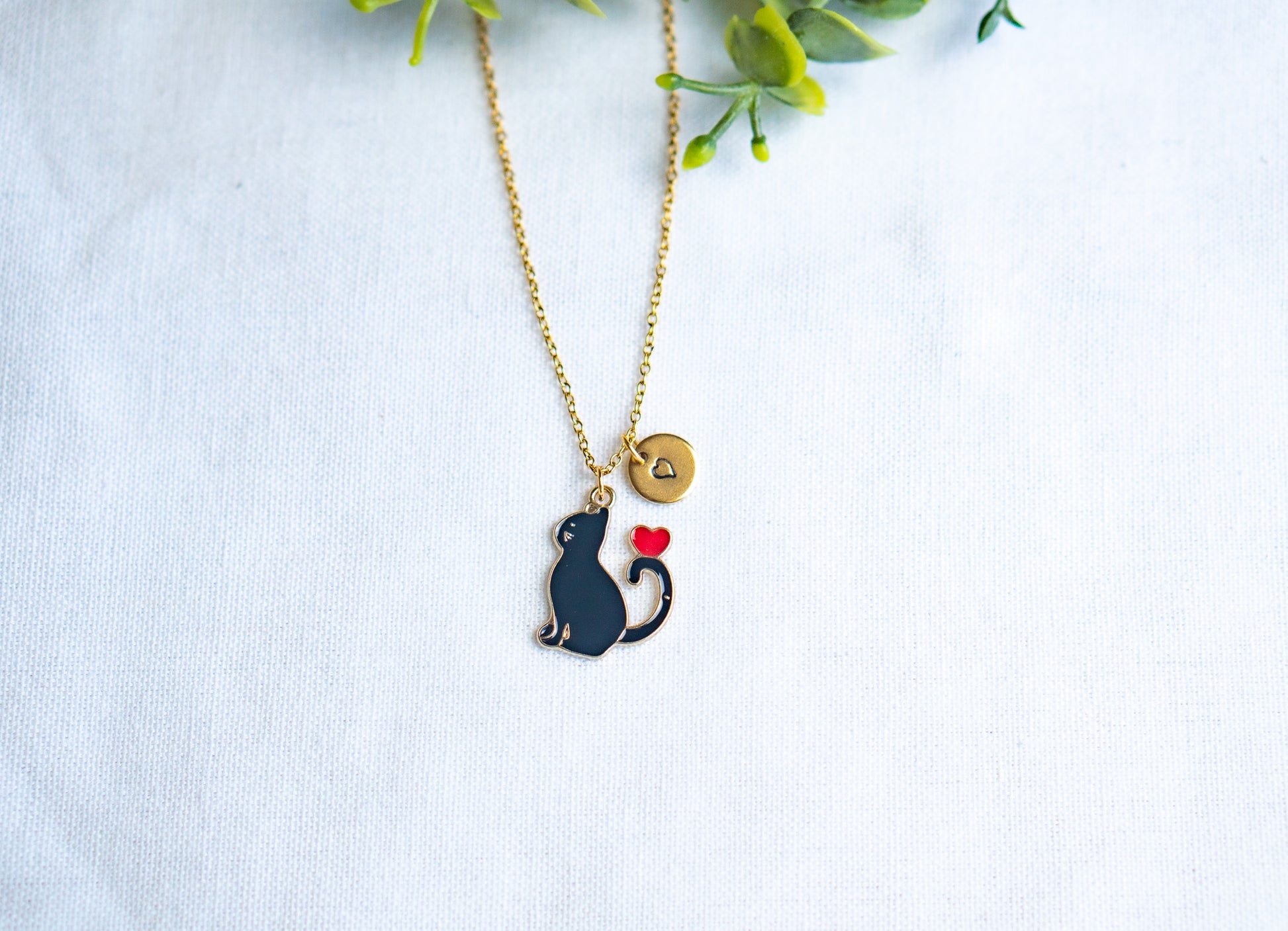 Close-up of a black cat-themed necklace with a red heart design on top of the cat’s black tail, displayed on a white fabric background.